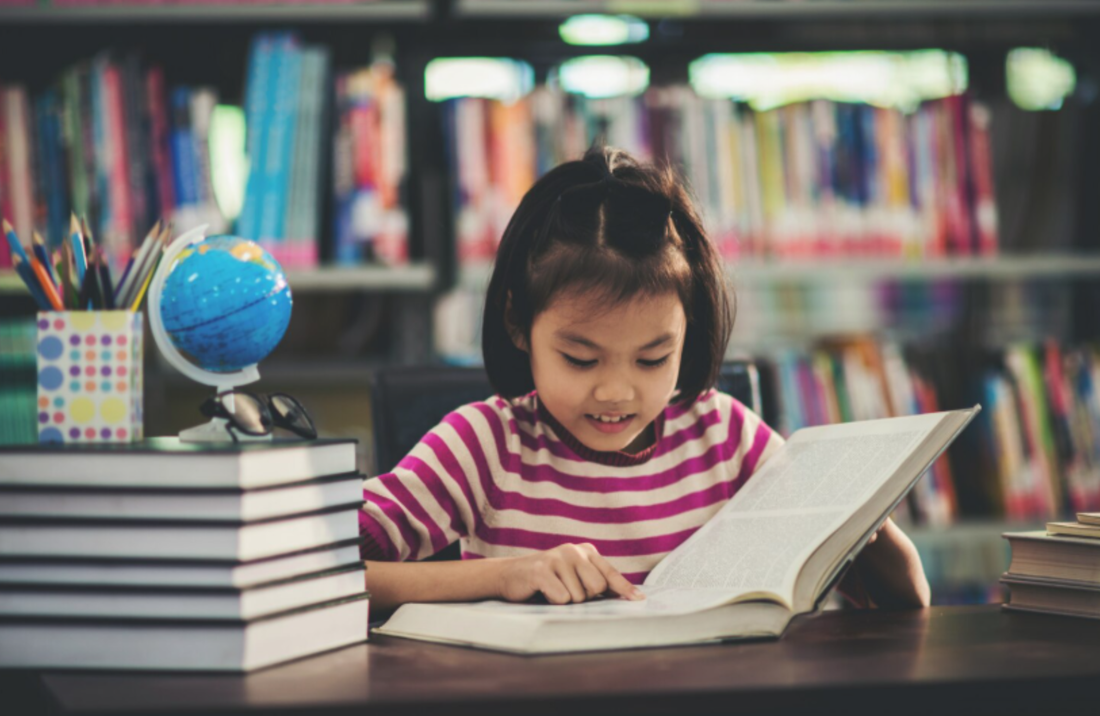 A young child sits at a table in a library, reading an open book with focused attention. Stacks of books, a small globe, and a cup of colored pencils are on the table, while bookshelves filled with books are visible in the background.